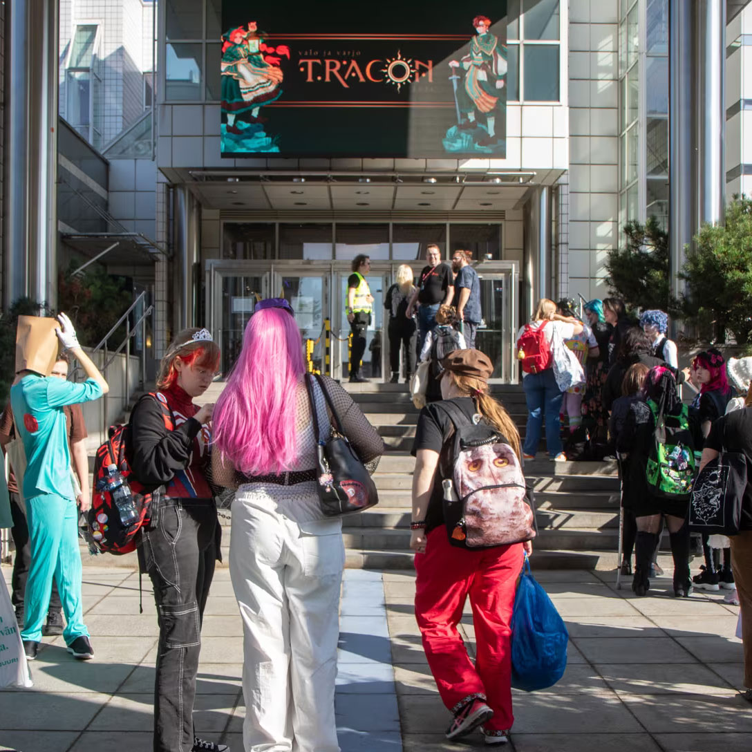 tracon_etusivu_yleiskuva_fapio_nelio Kuvan etualalla seisoo ja kävelee ihmisiä porrastasanteilla värikkäissä vaatteissa tai cosplayssa. He ovat osittain varjossa ja auringossa. Taustalla näkyy Tampere-talon pääovet. Ovien yllä olevassa näytössä on esillä Traconin vuoden 2024 teemakuvitusta.